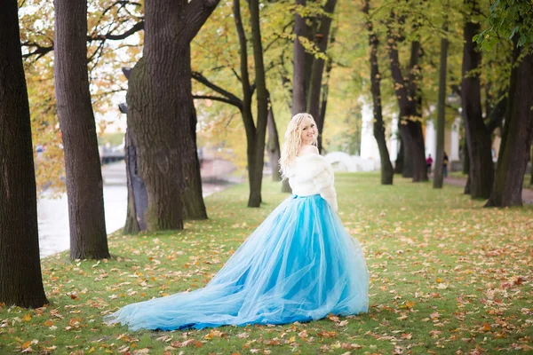 Blonde woman in sky blue fairy dress and white short fur coat walking in autumn park