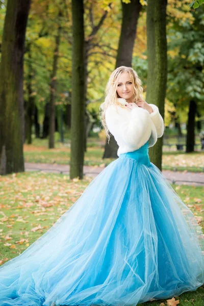 Blonde woman in sky blue fairy dress and white short fur coat walking in autumn park