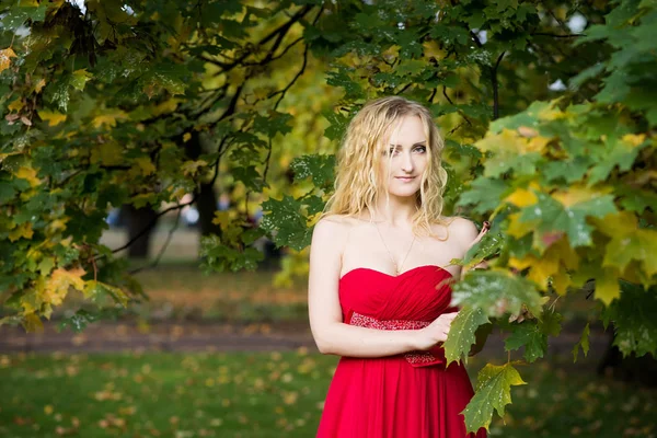 Blonde woman with wavy hair in satin red dress posing in autumn forest