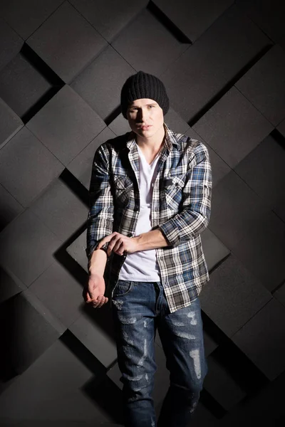 young hipster man in checked shirt and black hat posing on structural black background