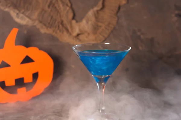 Bir cam kadeh mavi kokteyl ile Halloween kavramı. Duman etkisiyle. Karanlık bir arka planda. Arka planda bir kağıt kabak ile.