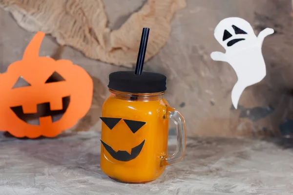 Halloween concept. Pumpkin or carrot drink in a glass jar on a gray textural background. With paper pumpkin and a ghost in the background.