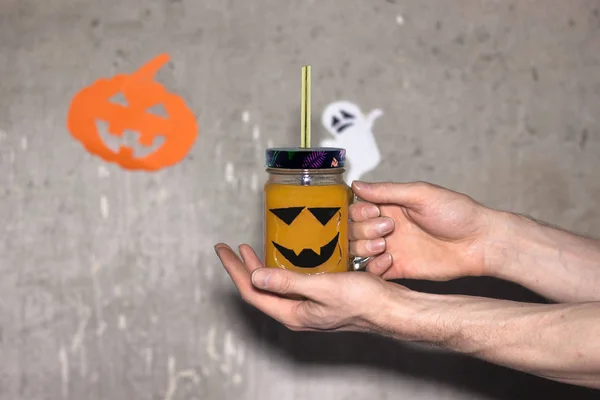 Halloween concept. Pumpkin or carrot drink in a glass jar. In men's hands. On a gray background. With paper pumpkin and ghost in the background.