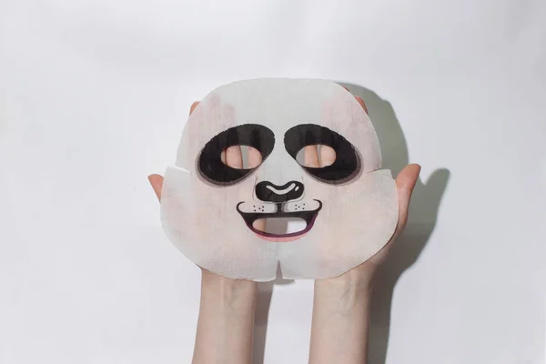 Sheet face mask. Fabric mask for facial skin. Female hands hold a fabric panda mask for the face. On a white background. View from above.Cosmetology, medicine and healthcare. The concept of home treatments for moisturizing the skin.