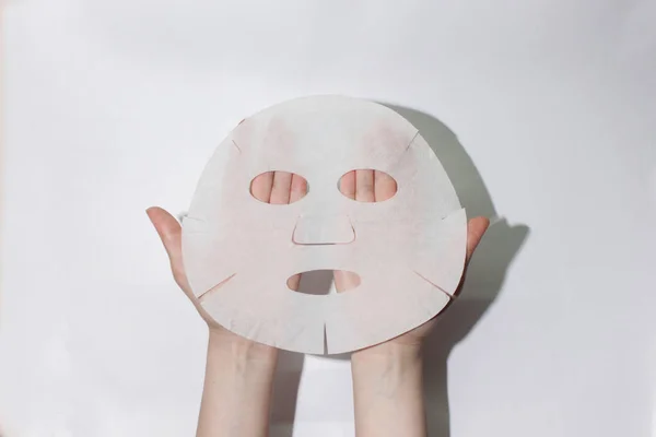 Sheet face mask. Fabric mask for facial skin. Female hands hold a face mask. On a white background. View from above.Cosmetology, medicine and healthcare. The concept of home treatments for moisturizing the skin.