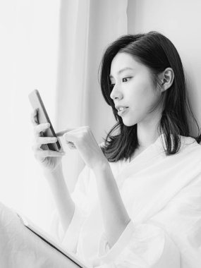 Genç Asyalı kadın evde siyah beyaz akıllı telefon kullanıyor.