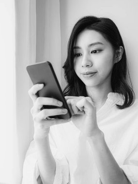 Genç Asyalı kadın evde siyah beyaz akıllı telefon kullanıyor.