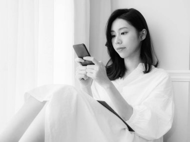 Genç Asyalı kadın evde siyah beyaz akıllı telefon kullanıyor.