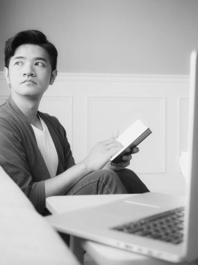 Genç Asyalı adam evde siyah beyaz dinlenirken kitap okuyor.
