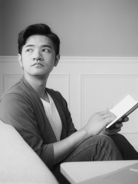 Genç Asyalı adam evde siyah beyaz dinlenirken kitap okuyor.