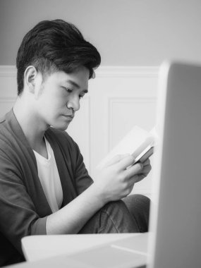 Genç Asyalı adam evde siyah beyaz dinlenirken kitap okuyor.