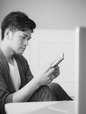 Genç Asyalı adam evde siyah beyaz dinlenirken kitap okuyor.