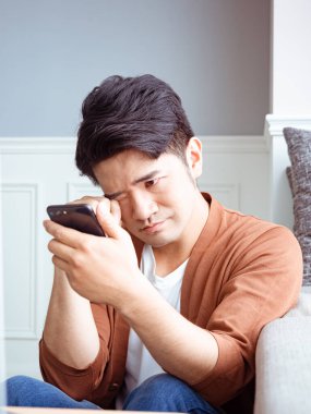 Asyalı genç adam yorgun gözlerini ovalıyor akıllı telefonunu..