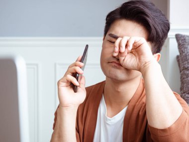 Asyalı genç adam yorgun gözlerini ovalıyor akıllı telefonunu..