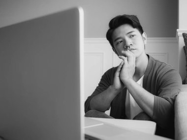 Asyalı genç adam dizüstü bilgisayarın önünde masada oturuyor.