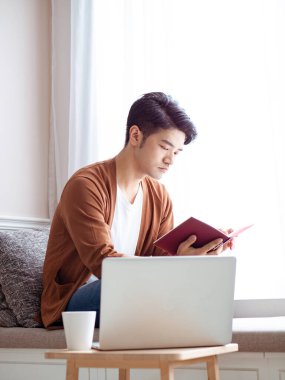 Asyalı genç adam dizüstü bilgisayarın önünde masada oturuyor.