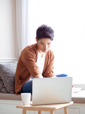 Asyalı genç adam dizüstü bilgisayarın önünde masada oturuyor.