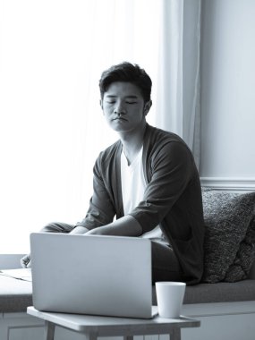 Asyalı genç adam dizüstü bilgisayarın önünde masada oturuyor.