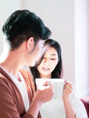 Genç Asyalı kadın ve erkek ellerinde bir fincan kahveyle evde vakit geçirmekten zevk alıyorlar..