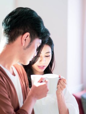 Genç Asyalı kadın ve erkek ellerinde bir fincan kahveyle evde vakit geçirmekten zevk alıyorlar..