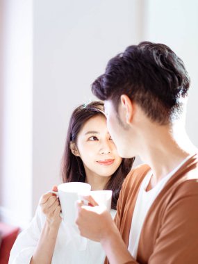 Genç Asyalı kadın ve erkek ellerinde bir fincan kahveyle evde vakit geçirmekten zevk alıyorlar..