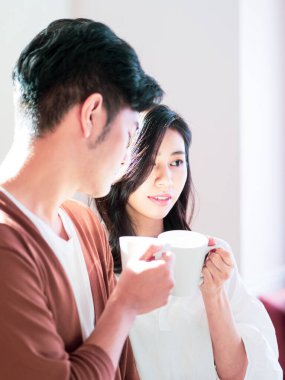 Genç Asyalı kadın ve erkek ellerinde bir fincan kahveyle evde vakit geçirmekten zevk alıyorlar..