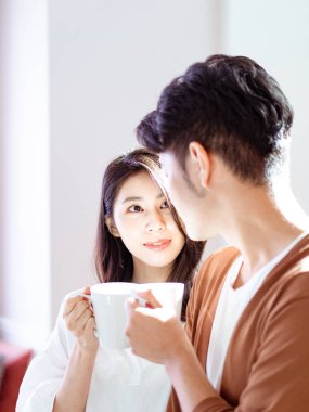 Genç Asyalı kadın ve erkek ellerinde bir fincan kahveyle evde vakit geçirmekten zevk alıyorlar..