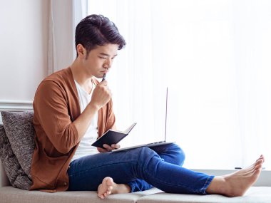 Genç Asyalı adam dizüstü bilgisayar kullanırken bir kitap yazıyor.