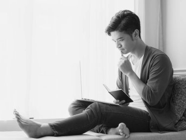 Genç Asyalı adam dizüstü bilgisayar kullanırken bir kitap yazıyor.