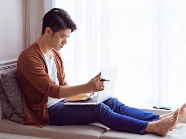 Genç Asyalı adam dizüstü bilgisayar kullanırken bir kitap yazıyor.