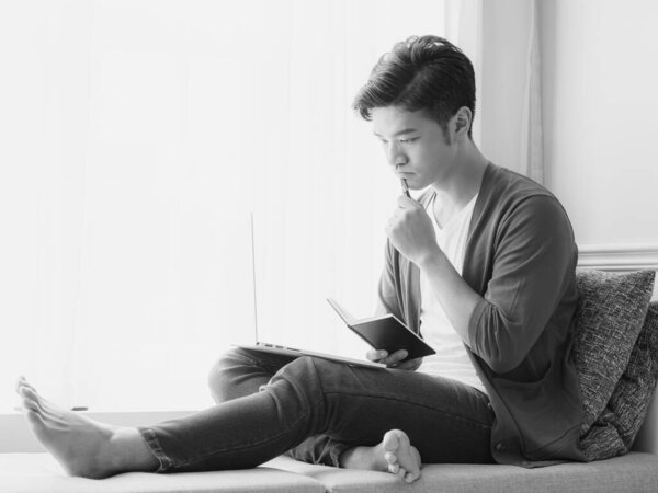 Young Asian man writing in a book when using laptop