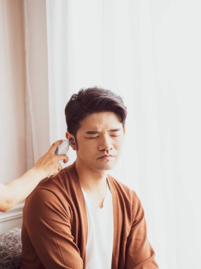 Genç Asyalı adam cerrahi maske takıyor ve kulak termometresi kullanıyor.