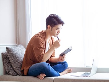 Genç Asyalı adam deftere yazı yazıyor ve dizüstü bilgisayar kullanıyor..