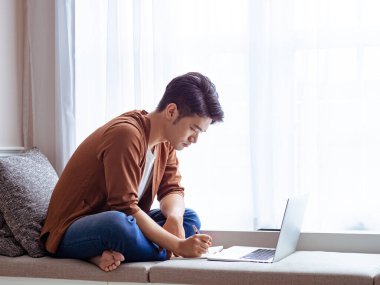 Genç Asyalı adam deftere yazı yazıyor ve dizüstü bilgisayar kullanıyor..