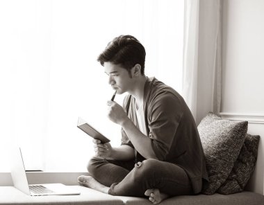 Genç Asyalı adam dizüstü bilgisayar kullanıyor ve deftere yazıyor..