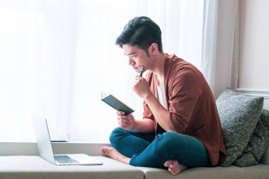 Genç Asyalı adam dizüstü bilgisayar kullanıyor ve deftere yazıyor..