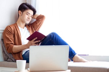 Genç Asyalı adam evde dinlenirken kitap okuyor.