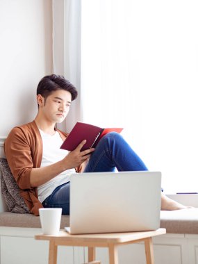 Genç Asyalı adam evde dinlenirken kitap okuyor.