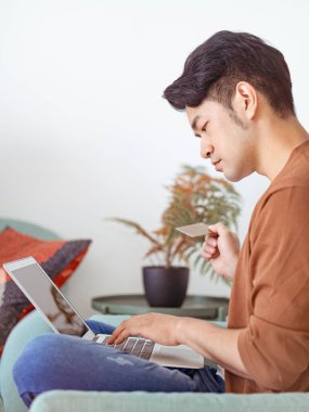 Genç adam evde kredi kartı ve laptop ile online alışveriş yapıyor.