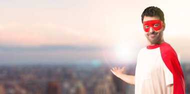 Süper kahraman maske ve kırmızı pelerin geri gün batımı bir arka plan üzerinde bir ürün sunan işaret parmağı ile işaret eden adamla