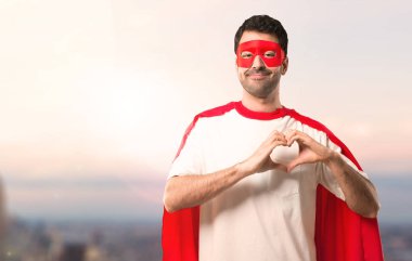Süper kahraman maske ve kırmızı pelerin eller tarafından kalp sembolü yapma adamla. Gün batımı bir arka plan üzerinde aşık olmak