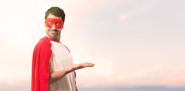 Süper kahraman maske ve kırmızı pelerin görünümlü doğru günbatımı bir arka plan üzerinde gülümseyen iken bir ürün ya da bir fikir sunmak adamla