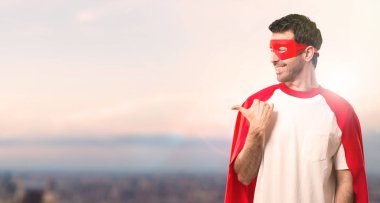 Bakarken bir ürün ya da bir fikir sunmak için maske ve kırmızı pelerin yan parmak ile işaret eden adamla süper kahraman ileri gün batımı bir arka plan üzerinde gülümseyen