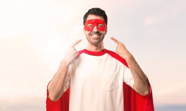 Süper kahraman maske ve kırmızı pelerin günbatımı bir arka plan üzerinde ağız ve yüz parmaklarıyla işaret ederken mutlu ve keyifli bir ifade ile gülümseyen adam