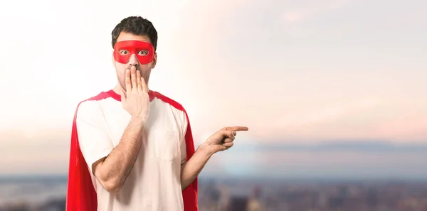 Superhero man with mask and red cape pointing finger to the side and presenting a product with a surprised face while covering mouth on a sunset background