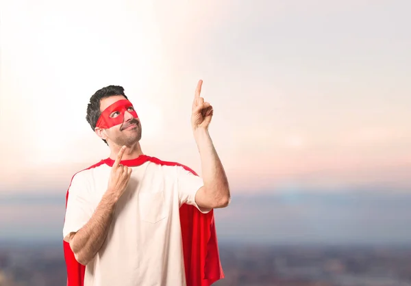 Superhero man with mask and red cape pointing with the index finger a great idea and looking up on a sunset background