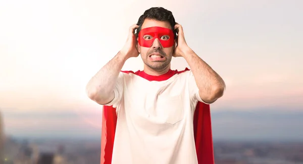 マスクと赤のケープ付きスーパー ヒーロー男は ための頭の上の手を取る片頭痛は 夕日を背景に — ストック写真
