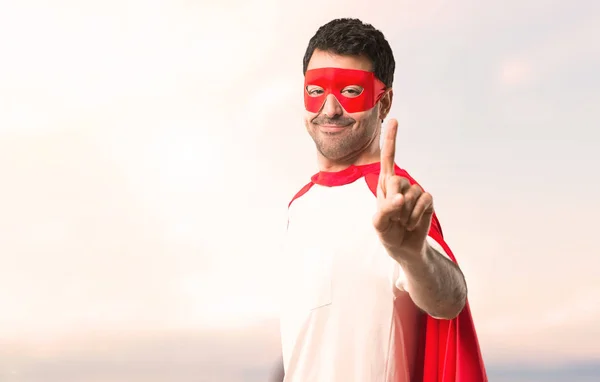 Superhero man with mask and red cape showing and lifting a finger in sign of the best on a sunset background