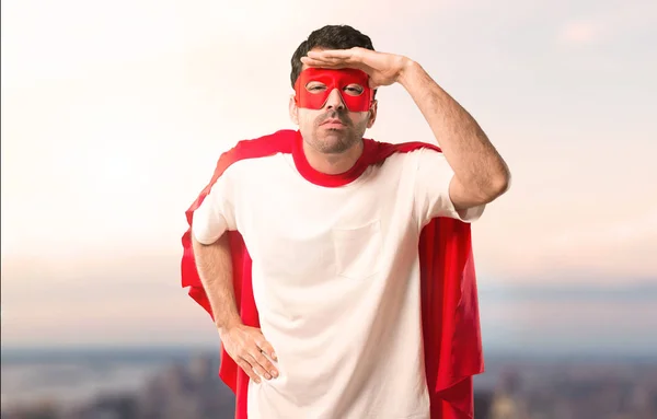 Superhero man with mask and red cape looking far away with hand to look something on a sunset background