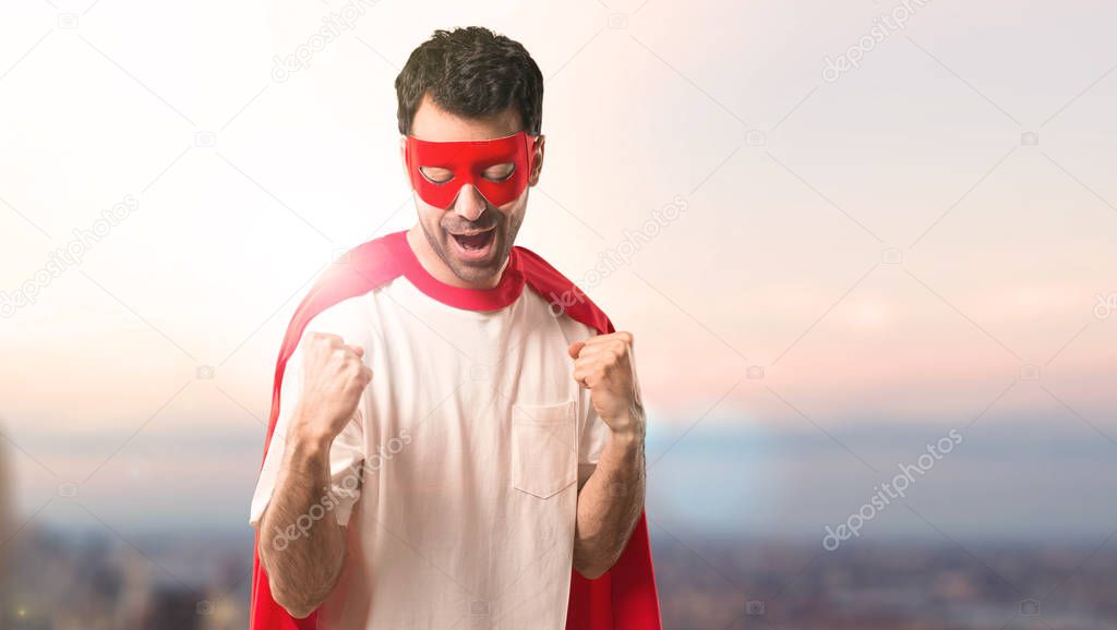 Superhero man with mask and red cape celebrating a victory and happy for having won a prize on a sunset background